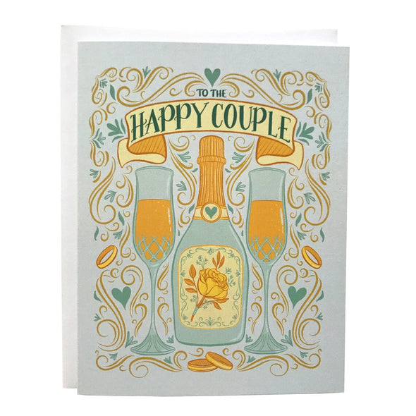 A pastel blue greeting card features an illustration of two full champagne flutes, a champagne bottle with a yellow rose, wedding bands, Art Nouveau flourishes, hearts, and the words "To The Happy Couple". The card sits against a white envelope on a white background.