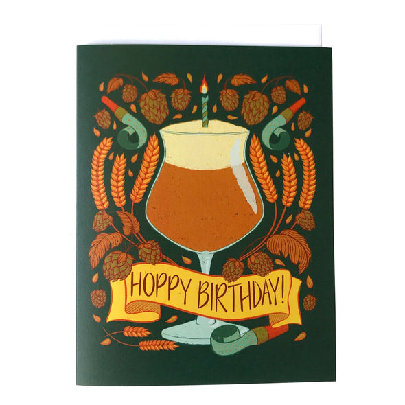 A dark green greeting card features an illustration of a full and frothy beer glass with a birthday candle in it. At the bottom of the glass, a yellow banner reads "Hoppy Birthday". Around the glass are stalks of wheat, wheat berries, hops blossoms, and light blue noisemakers. The card sits against a white envelope on a white background.