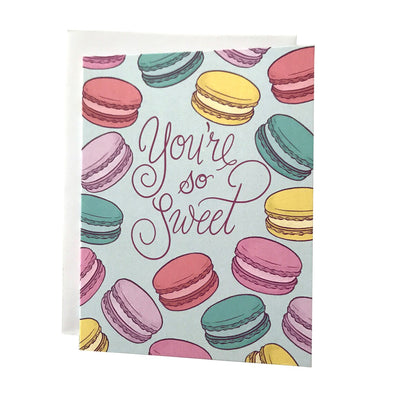 A pastel greeting card features the words "You're so Sweet" in purple letters amid a scattered array of purple, pink, yellow and blue macarons. The card sits against a white envelope on a white background.